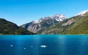 Stunning view of a turquoise lake surrounded by snow-capped mountains in Alaska under a clear blue sky.