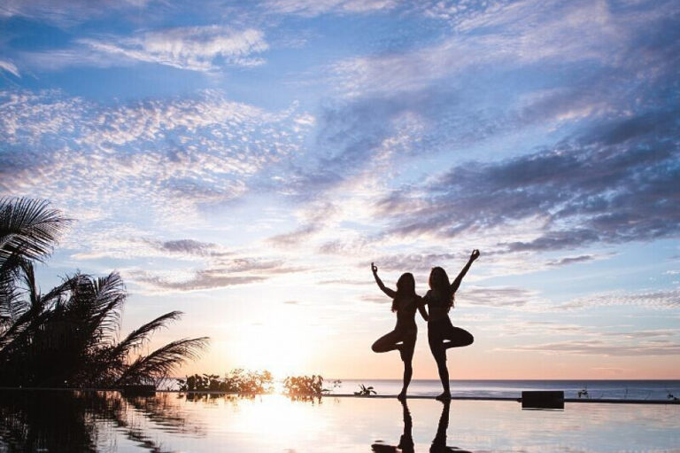 Two women practicing yoga at sunset by pool