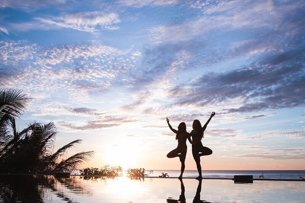 Two women practicing yoga at sunset by pool