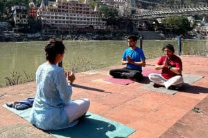 Three people meditating by riverside with bridge backdrop