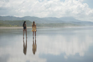Two women standing on reflective beach shoreline