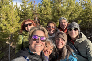 Six women smiling in pine forest selfie